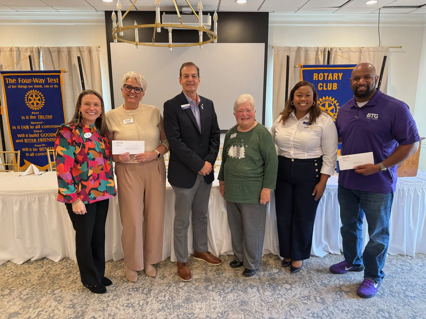 Six people standing together at a Rotary Club of Newnan meeting, two of them holding grant check envelopes, in front of the club&rsquo;s Four-Way Test and Rotary Club banners.