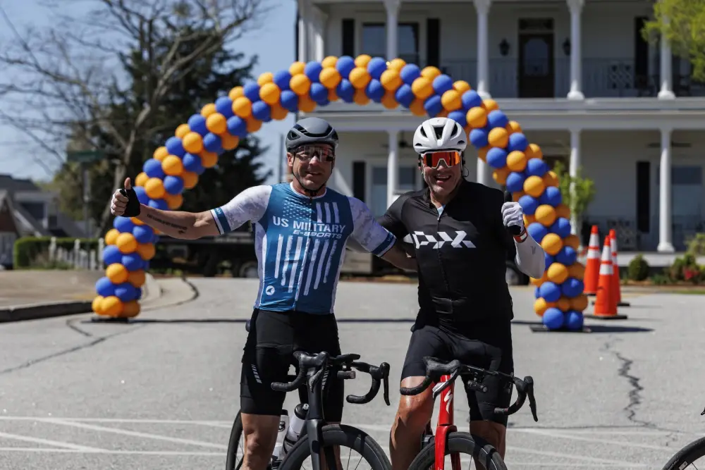 Two riders stand with their bicycles under a blue and gold balloon arch at the finish, thumbs up, in front of a white columned house.
