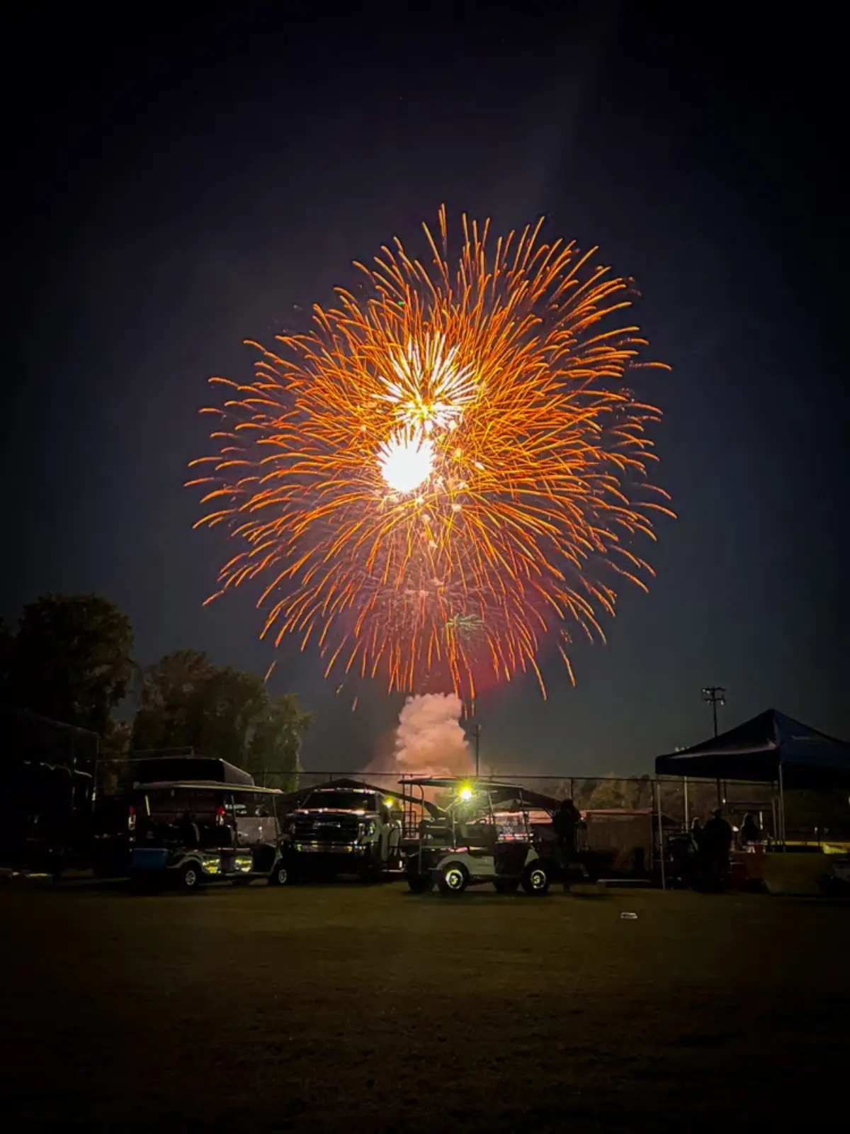 A large gold firework bursting over the field at Fireworks Over Newnan, with golf carts and tents silhouetted below.
