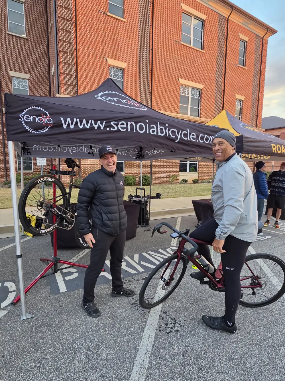 Two men laughing beside Senoia Bicycle&rsquo;s sponsor tent at the start line, one holding a road bike, with a repair stand behind them.