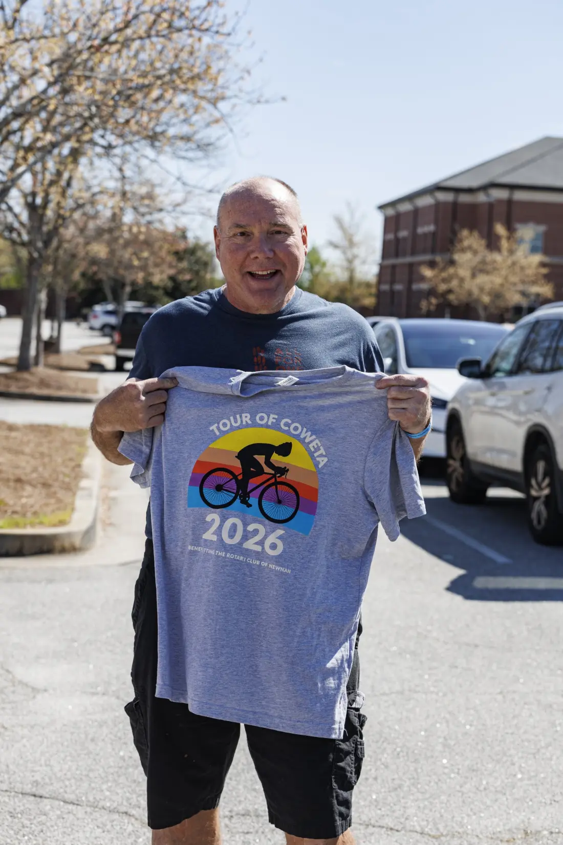 A rider holding up the gray 2026 Tour of Coweta event shirt, printed with a cyclist silhouette against a rainbow sun and the words benefitting the Rotary Club of Newnan.